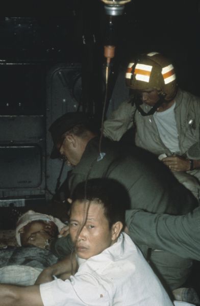 U.S. helicopter evacuation of wounded Vietnamese fighter to hospital  including treatment by U.S. service medics and corpsmen. A man with a bandaged head is lying on a stretcher while three men bend over him.  An IV is hanging from the ceiling in the metal interior.