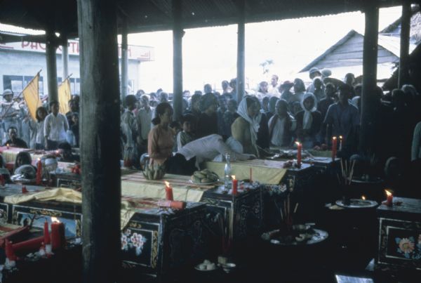 Large funeral for multiple people in Binh Hung, Vietnam. Numerous decoratively painted coffins with cloth coverings are on display in a large open-air, roofed structure. On top of the coffins ae bunches of fruit, red candles and sticks of incense. A large crowd looks on from outside, and a few people are standing or crouching near the individual coffins.