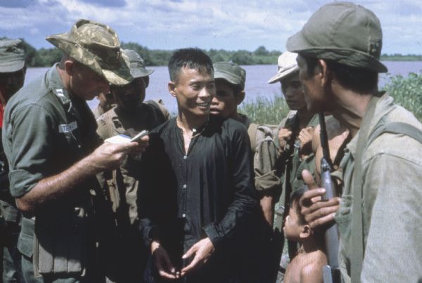 An American soldier inspects the papers of a Vietnamese man in the countryside along a river. Vietnamese soldiers and a boy are grouped around him.