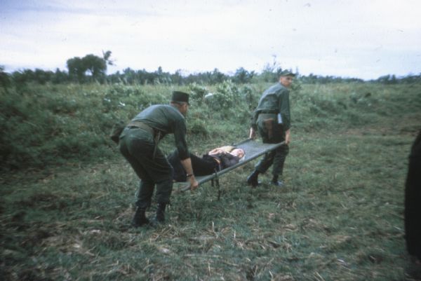 Two American soldiers carry a Vietnamese woman on a stretcher across a field in Vietnam.