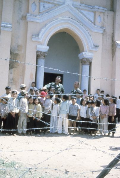 View through barbed wire fence of a group of about 35 school-aged Vietnamese children standing around two American soldiers in front of a large peach and white building in Vietnam. One of the soldiers is putting his hat on one of the children.