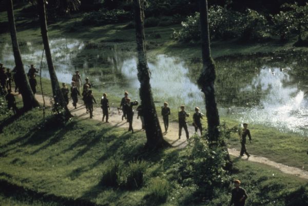 Elevated view of a group of Vietnamese soldiers wearing berets hiking through a forest in Vietnam. The footpath they are on is bordered on one side by trees and on the other with a marshy expanse of water and plants.