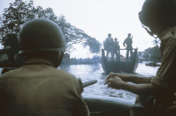 Sea Swallows, an anti-communist Vietnamese militia, begin an operation by boats on a canal in the vicinity of Van Dinh, Vietnam. Militia members hold guns and wear helmets in a boat in the foreground. Another boat is ahead of them.