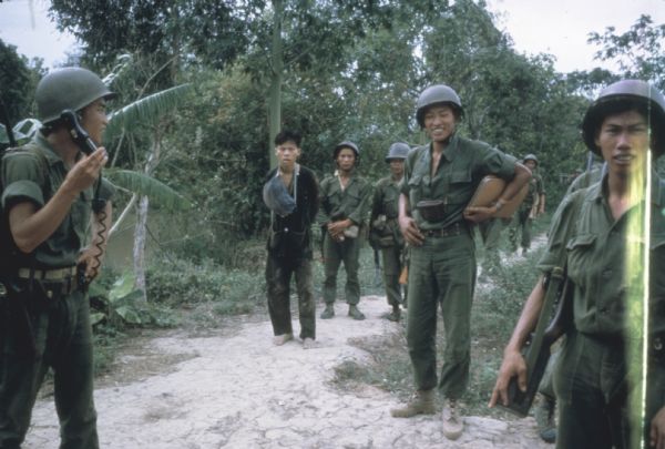 Small group of Vietnamese soldiers stand with a recently captured prisoner suspected of being Viet Cong on a path in the woods in Vietnam. One soldier is using a field radio and the barefoot prisoner is bound, his black outfit is muddy, and a satchel is hanging around his neck.