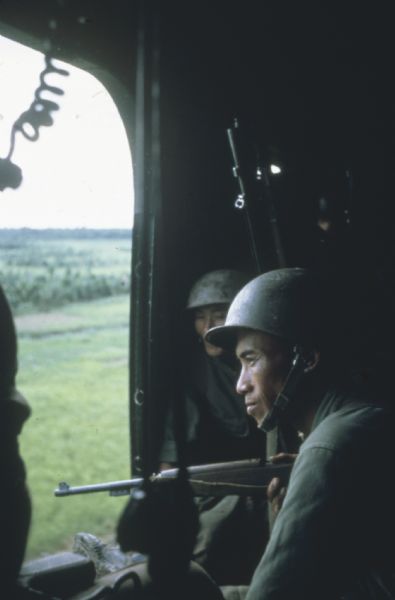 Vietnamese soldier in a helicopter, holding a rifle and looking out the open door at the countryside of Vietnam.