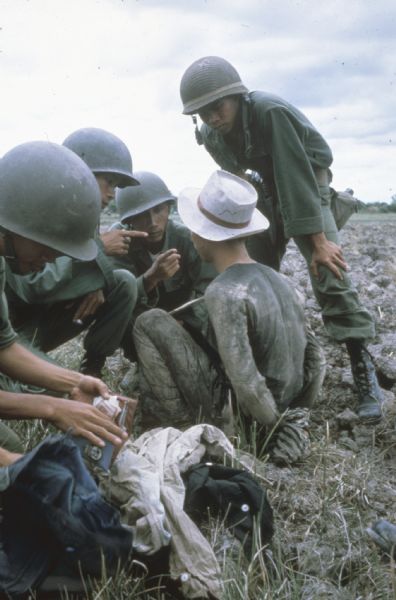 A small group of Vietnamese soldiers question a prisoner in a dry and rocky field in Vietnam. The soldiers are standing or crouching very close to the bound and muddy seated prisoner and pointing in his face. Another soldier searches the prisoner's bag and wallet.