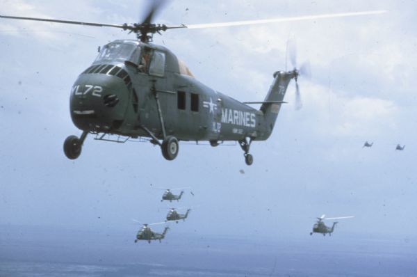 Aerial view of flock of U.S. Marine helicopters in the sky over Vietnam. One of the dark green and yellow helicopters is in the foreground. Four others trail close behind.