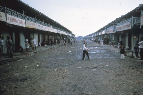 Main street in a Vietnamese town. A woman carrying a baby is crossing the unpaved and puddle-dotted street. Other adults and children are standing on the sides on the sidewalks. Balconies with business signs overhang the sidewalks.