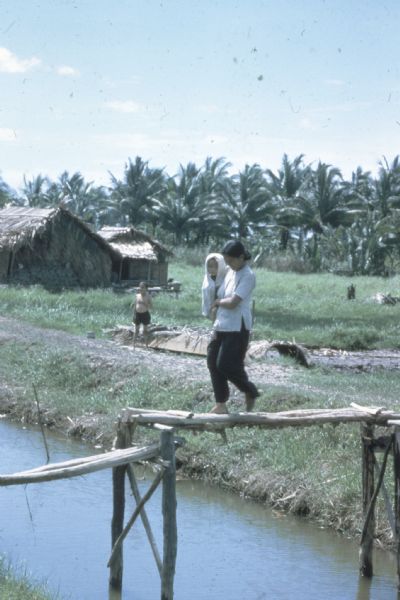 A woman carrying a child wrapped in a cloth crosses a log footbridge over a canal in the countryside in Vietnam. A boy is standing in the background and there are also two thatched structures.