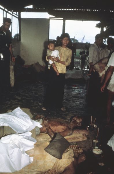 A woman holding a baby stands in front of two dead bodies laid out for a funeral in a open-air, covered building. The men were killed during a Viet Cong attack on the village of Vinh Quoi, Vietnam. An American soldier watches her from the edge of the frame and other Vietnamese adults and children are also standing around in the room.