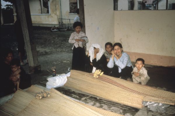 Women and children crouch around bodies laid out for funerals covered with woven mats. The deceased were killed during a Viet Cong raid of Vinh Quoi, Vietnam. The arrangements for the dead feature sticks of incense and bunches of bananas. One of the women is crying and one is covering her head with a cloth. A girl standing next to them has an injured face.
