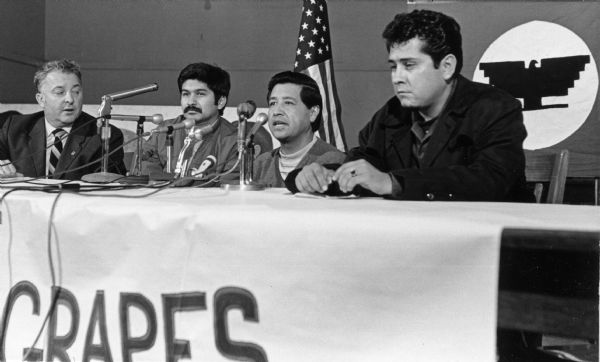 John Schmitt, AFL CIO president; Manuel Salas, labor contractor for Libby, McNeil and Libby; Cesar Chavez and Eliseo Medina sit together on a panel in Milwaukee speaking about the grape boycott. The table has numerous microphones and has a banner hanging on it that reads in part "Grapes." An American flag and an Farm Workers AFL-CIO flag are hanging in the background.
<p></p>Panel del boicot de uvas en Milwaukee
<p></p>John Schmitt, presidente de AFL CIO; Manuel Salas, contratante laboral para Libby, McNeil & Libby; Cesar Chávez y Eliseo Medina sentados durante un panel en Milwaukee para hablar acerca del boicot de uva. La mesas tienen micrófonos numerosos y un estandarte colgando que en parte se lee la palabra "Uvas" ("Grapes"). Una bandera americana y una bandera de Farm Workers AFL-CIO se pueden ver colgando en el fondo.