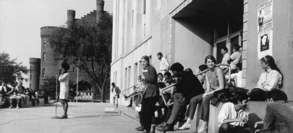 Protest on the Library Mall at the University of Wisconsin-Madison in support of the grape boycott. A woman stands on the sidewalk speaking at a microphone. Students sit and stand clustered on the steps and walls. The Red Gym and a large tree are visible in the background. There is a poster on Memorial Library announcing a no work, no school strike in response to the kidnapping of Bobby Seale.
<p></p>Protesta apoyando el boicot de uvas en la University of Wisconsin-Madison
<p></p>Protesta en Library Mall de la University of Wisconsin-Madison para apoyar el boicot de uva. Una mujer está parada en la acera y hablando con un micrófono. Hay estudiantes sentados y parados alrededor de los escalones y las paredes. El edificio Red Gym y un árbol grande están visibles hacia el fondo. Hay un póster en la biblioteca Memorial Library anunciando una huelga de no trabajo y no escuela en respuesta al secuestro de Bobby Seale.