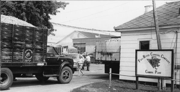 Entrance to Fall River Canning Company. A number of trucks full of vegetables are in the parking lot or driveway in front of low sprawling warehouse buildings. A man and a woman are in the driveway.
<p></p>Entrada de la compañía Fall River Canning Company
<p></p>Entrada de la compañía enlatadora Fall River Canning Company. Unos camiones llenos de vegetales están en el estacionamiento o camino de entrada enfrente de unos edificios de almacén. Un hombre y una mujer están en el camino de entrada.