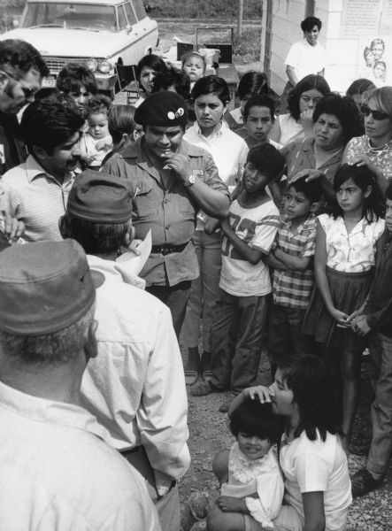 Jorge Vasquez (wearing beret) discusses strategy with Fall River Canning Company workers after walkout. Children, youth and adults stand clustered in a circle around Vasquez in a gravel parking lot.
<p></p>Trabajadores discutiendo una estrategia después de un paro laboral
<p></p>Jorge Vásquez (vistiendo una boina) discute estrategia con trabajadores de Fall River Canning Company después de un paro laboral. Niños, adolescentes, y adultos están parados en un círculo alrededor de Vásquez en un estacionamiento de grava.