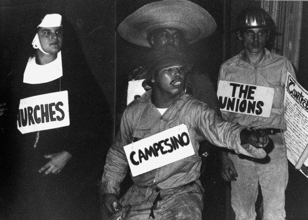 Teatro Campesino acto or skit dramatizing the experiences of Hispanic workers in America. Four actors stand together in costume with signs around their necks identifying themselves. The "Campesino" stands in front pointing at the audience. In the background stand "Churches," "The Unions" and another character whose sign is obscured.
<p></p>
In the background and wearing a sombrero is Luis Valdez, playwright  filmmaker, and founder of Teatro Campesino.

<p></p>Acto de Teatro Campesino
<p></p>Un acto de Teatro Campesino dramatiza las experiencias de los trabajadores hispanos en América. Cuatro actores parados juntos, en disfraz, y con letreros colgando de sus cuellos que los identifica. El "Campesino" está parado enfrente y señalando a la audiencia. Hacia el fondo están "las iglesias"/"Churches," "los sindicatos"/"The Unions," y otro personaje con un letrero que no se puede ver. Hacia el fondo y con un sombrero está Luis Valdez, dramaturgo cineasta y fundador de Teatro Campesino.

