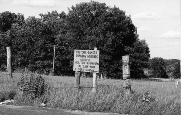 Dump where discarded mattresses were resold to growers for migrant laborer camp use. A sign identifying the dump and its hours stands behind a barbed wire fence in a grassy area, in front of a screen of trees.
<p></p>Basurero de Wautoma-Dakota
<p></p>Basurero donde colchones tirados eran re-vendidos a los agricultores para usar en los campos de trabajadores emigrantes. Un letrero, identificando el basurero y sus horas de operación, está detrás de una cerca de alambre en una área con pasto y enfrente de una cubierta de arboles.