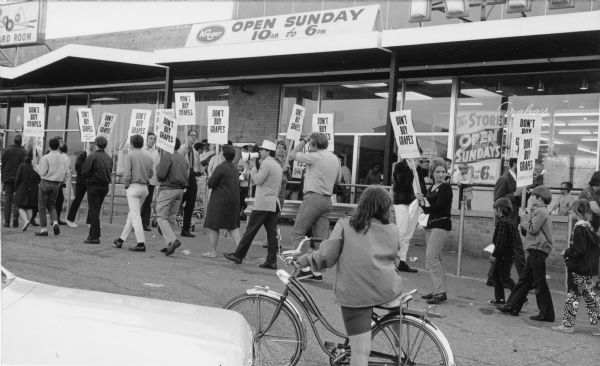 A group of picketers supporting the nationwide United Farmworkers/AFL-CIO grape boycott in front of Kroger Supermarket. Picketers are carrying signs that say "Don't Buy Grapes." At the center is a man wearing sunglasses and a hat walking and speaking through a bullhorn. Among picketers are three young children on the right. A girl wearing a jacket and shorts, with her back towards the camera, observes the event on her bicycle while standing in front of a parked car. Grape boycotts in Wisconsin were organized by the local farm-worker labor union, Obreros Unidos (United Workers), an independent farm worker labor union effort in the 1960s.<p>Manifestantes de Milwaukee apoyando el boicot de uvas<p>Un grupo de manifestantes apoyando el boicot de uvas nacional de United Farmworkers/AFL-CIO enfrente del supermercado Kroger Supermarket. Manifestantes están sosteniendo letreros que dicen "No compren uvas" ("Don't Buy Grapes"). Al centro está un hombre con lentes de sol y una gorra, caminando y hablando con un megáfono. Entre los manifestantes hay tres niños hacia la derecha. Una niña vistiendo una chaqueta y un pantalón corto, con su espalda hacia la cámara, observa el evento en su bicicleta mientras está parada enfrente de un carro estacionado. Los boicots de uva en Wisconsin fueron organizados por el sindicato local de trabajadores agrícolas, Obreros Unidos, un esfuerzo independiente del sindicato de trabajadores agrícolas durante los años 1960.</p>