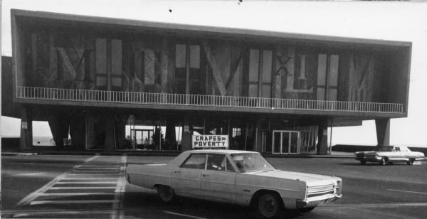 A sign on the roof of a car that says, "Grapes = Poverty" supporting the United Farmworkers (UFW)/AFL-CIO grape boycott. The car is parked on a street in front of the Milwaukee County War Memorial. The nationwide UFW/AFL-CIO grape boycott movement to improve conditions for migrant farm workers was started in California by activist Cesar Chavez. Inspired, the Wisconsin farm-worker labor union, Obreros Unidos (United Workers) organized boycott action in every major Wisconsin city.<p>Un carro con un letrero del boicot de uvas<p>Un letrero en el techo de un carro que dice "Uvas = Pobreza" ("Grapes = Poverty") para apoyar el boicot de uvas de United Farmworkers (UFW)/AFL-CIO. El carro está estacionado en una calle enfrente de Milwaukee County War Memorial (monumento a los caídos en la guerra). El movimiento nacional del boicot de uvas organizado por UFW/AFL-CIO para mejorar las condiciones de los trabajadores agrícolas emigrantes fue empezado en California por el activista Cesar Chávez. Obreros Unidos, el sindicato de trabajadores agrícolas en Wisconsin, fue inspirado a organizar acciones de boicot en cada ciudad mayor de Wisconsin.</p>