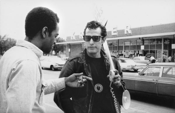 Ernesto Chacon, a grape boycott organizer from Milwaukee, is wearing a jacket, sunglasses and a necklace around his neck. He is holding a United Farmworkers/AFL-CIO grape boycott sign that says, "Don't Buy Grapes" and a bullhorn in the parking lot of Kroger Supermarket. He is facing Bill Smith, a union organizer with Obreros Unidos,  on the left, who is pointing towards the bullhorn. Grape boycotts in Wisconsin were organized by the local farm-worker labor union, Obreros Unidos (United Workers) an independent farm worker labor union effort in the 1960s.<p>Piquete del boicot de uvas
<p>Ernesto Chacón, un organizador del boicot de uva en Milwaukee, está vestido en una chaqueta, con lentes de sol, y un collar alrededor de su cuello. Está sosteniendo un letrero de United Farmworkers/AFL-CIO para el boicot de uvas que dice "No Compren Uvas" ("Don't Buy Grapes") y un megáfono mientras en el estacionamiento del supermercado Kroger Supermarket. Está cara a cara con Bill Smith, un organizador sindicalista con Obreros Unidos, que está a la izquierda y señalando al megáfono. Los boicots de uva en Wisconsin fueron organizados por el sindicato local de trabajadores agrícolas, Obreros Unidos, un esfuerzo independiente del sindicato de trabajadores agrícolas durante los años 1960.</p>