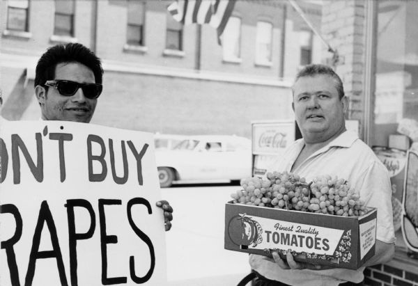 On the left, Ernesto Chacon, is in sunglasses and holding a sign that says, "Don't Buy Grapes," picketing for the United Farmworkers/AFL-CIO grape boycott. Grape boycotts in Wisconsin were organized by the local farm-worker labor union, Obreros Unidos (United Workers),an independent farm worker labor union effort in the 1960s. The man on the right is holding a tomato box filled with grapes, possibly counter-picketing the grape boycott in front of a store.<p>Haciendo piquete por el boicot de uvas<p>A la derecha, Ernesto Chacón con lentes de sol y sosteniendo un letrero que dice "No Compren Uvas" ("Don't Buy Grapes"), mientras hace piquete durante el boicot de uvas de United Farmworkers/AFL-CIO. Los boicots de uva en Wisconsin fueron organizados por el sindicato local de trabajadores agrícolas, Obreros Unidos, un esfuerzo independiente del sindicato de trabajadores agrícolas durante los años 1960. El hombre a la derecha está sosteniendo una caja para jitomates llena de uvas, posiblemente haciendo contra-piquete (protestando) el boicot de uva enfrente de la tienda.</p>