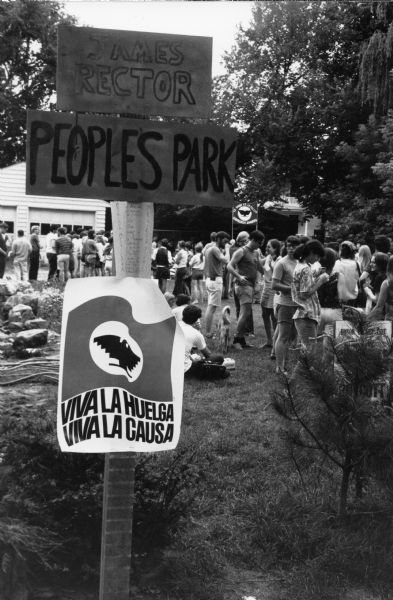 A benefit gathering at James Rector People's Park to support the United Farmworkers (UFW) grape boycott. The UFW sign, with its eagle symbol,  posted in the foreground says, "Viva la Huelga/Viva la Causa" (Long Live the Strike/Long Live the Cause). Above it is a sign that says, "Comida Mexicana" (Mexican Dinner). In the background, young people are partaking in the benefit and a UFW sign stands with the message, "La Lucha es la Fuerza/ Obreros Unidos" (The Struggle is the Strength - United Workers).<p>Grape boycotts in Wisconsin were organized by the local farm-worker labor union, Obreros Unidos (United Workers), an independent farm worker labor union effort in the 1960s.<p>Beneficio en Madison para UFW en el People’s Park<p>Una reunión beneficiaria en el James Rector People's Park para apoyar el boicot de uvas de United Farmworkers (UFW). El letrero de UFW con el símbolo de una águila está hacia el fondo y dice "Viva la Huelga/Viva la Causa." Arriba hay otro letrero que dice "Comida Mexicana." Hacia el fondo hay gente joven participando en el beneficio y un letrero de UFW que dice "La Lucha es la Fuerza/Obreros Unidos."<p>Los boicots de uva en Wisconsin fueron organizados por Obreros Unidos, un esfuerzo independiente del sindicato de trabajadores agrícolas durante los años 1960.</p>