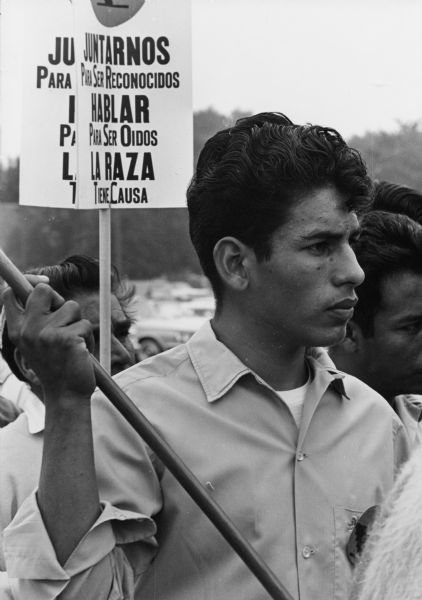 An Obreros Unidos (United Workers) member holding a sign, possibly like those in the background, that says "Juntarnos Para Ser Reconocidos/Hablar Para Ser Oidos/La Raza Tiene Causa" which translates to "Join To Be Recognized/Speak to Be Heard/The People Have A Cause." On August 15, 1966, 24 marchers left Wautoma, Wisconsin, headed west to Coloma, then south along State Highway 51 and reached the Capitol in Madison after five days and eighty miles of walking. Led by Jesús Salas, marchers went to state agencies to present the following demands: $1.25 minimum wage; better housing; insurance for accidents and hospitalization; public toilet facilities for farm worker use; and a meeting with the Governor's Committee on Migratory Labor.<p>Obreros Unidos grew in Wisconsin during the 1960s and had deep roots in South Texas and Mexico. It was formed to deal with discrimination and oppression migrant farm workers were facing while working and living in  Wisconsin and elsewhere along the journey to the North. In 1946, more than 4,000 farmworkers from Texas migrated to Wisconsin to cultivate and harvest vegetable and fruit crops. In 1961,85 percent of about 18,000 migrant workers, including 5,000 children under age 16, were recruited from Texas to work in Wisconsin.<p>Obreros Unidos marchan hacia Madison<p>Un miembro de Obreros Unidos sosteniendo un letrero, posiblemente uno como otros en el fondo, que dice "Juntarnos Para Ser Reconocidos/Hablar Para Ser Oídos/La Raza Tiene Causa" y se traduce a "Join To Be Recognized/Speak to Be Heard/The People Have A Cause" en inglés. El 15 de agosto de 1966, 24 manifestantes se fueron de Wautoma, Wisconsin, siguiendo al oeste hacia Coloma, luego al sur a lo largo de la carretera estatal State Highway 51, y llegaron al Capitolio en Madison después de cinco días y ochenta millas caminando. Dirigidos por Jesús Salas, los manifestantes visitaron agencias estatales para presentar las siguientes demandas: un sueldo mínimo de $1.25 por hora; mejores viviendas; seguro de accidentes y hospitalización; baños públicos para uso de los trabajadores; y una junta con el Governor's Committee on Migratory Labor (comité del gobernador que trata con la fuerza laboral migratoria).<p>Obreros Unidos se desarrollo en Wisconsin durante los años 1960 y tenía raíces profundas en el sur de Texas y México. Fue fundado para combatir la discriminación y opresión que los trabajadores agrícolas emigrantes estaban sufriendo mientras trabajaban y vivían en Wisconsin y en otros lugares a lo largo de su viaje al norte. En 1946, más de 4,000 trabajadores agrícolas de Texas emigraron hacia Wisconsin para cultivar y cosechar vegetales y fruta. En 1961, 85 por ciento de aproximadamente 18,000 trabajadores emigrantes, incluyendo 5,000 niños menores de 16 años, fueron reclutados de Texas para trabajar en Wisconsin.</p>