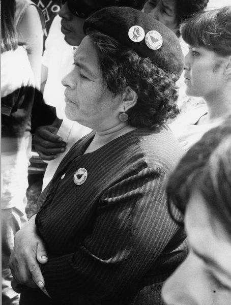 A Huelgusta (striker) against Fall River Canning Company with Obreros Unidos (United Workers), standing with a crowd. She is wearing round earrings, a black pin-striped jacket and a black beret. Pinned on her jacket is a United Farmerworkers (UFW) button with its eagle symbol and the message, "Viva la Causa" (Long Live the Cause)/Solidarity Forever." On her beret are two buttons, with the same button on the right and a button with a clenched fist on the left.  Grape boycotts in Wisconsin were organized by the local farm-worker labor union, Obreros Unidos, an independent farm worker labor union effort in the 1960s.<p>Huelguista en Cambria<p>Una huelguista contra la compañía Fall River Canning Company con Obreros Unidos, está parada entre una multitud. Está vistiendo aretes redondos, una chaqueta negra con rayas, y una boina negra. En su chaqueta trae un botón de United Farmerworkers (UFW) con el símbolo de la águila y el mensaje "Viva la Causa"/"Solidaridad para Siempre " ("Solidarity Forever"). Tiene dos botones en su boina, con el mismo mensaje en el botón a la derecha y el botón a la izquierda con la imagen de un puño. Las huelgas en Wisconsin fueron organizadas por Obreros Unidos, un esfuerzo independiente del sindicato de trabajadores agrícolas durante los años 1960.</p>