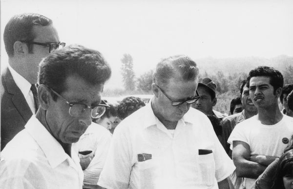 Fall River Canning Company's president William Evans stands in the center with migrant farm workers around him after their walkout.<p>Eighty workers of Hispanic descent had walked off their jobs charging the Fall River Canning Company with racially discriminatory practices. This labor action was a part of the statewide protest movements of the Obreros Unidos (United Workers), a union created to fight for migrant farm worker protection. In the background, wearing a dark suit is Evans' attorney.<p>William Evans el presidente de Fall River Canning Company está parado al centro con trabajadores agrícolas emigrantes a su alrededor después de que ellos hicieron un paro laboral.<p>Ochenta trabajadores de origen hispano dejaron su trabajo en acusación de prácticas racialmente discriminatorias por parte de Fall River Canning Company. Esta acción laboral fue parte de movimientos de protesta en todo el estado por parte de Obreros Unidos, un sindicato creado para luchar por la protección de los trabajadores agrícolas emigrantes. Hacia el fondo, vistiendo un traje oscuro está el abogado de Evans.</p>