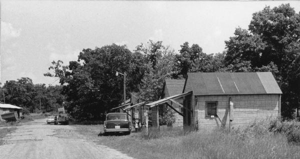 A central Wisconsin labor camp consisting of a row of wooden framed building with asphalt siding which housed migrant laborers. At the center, three children are standing in front of a building beside a black car looking into the camera. Farther down the road on the left is a shack and a worker standing by a car across from it.<p>In 1946, more than 4,000 farmworkers from Texas migrated to Wisconsin to cultivate and harvest vegetable and fruit crops. In 1961,85 percent of about 18,000 migrant workers, including 5,000 children under age 16, were recruited from Texas to work in Wisconsin.<p>In 1942-1964, more than 4 million Mexican workers entered the U.S. for temporary employment under the Bracero Program which was an agreement between the U.S. and Mexico. The agreement was created in response to a lobby from growers in western states who anticipated labor shortages during World War II and the Korean War.<p>Campo laboral en el centro de Wisconsin<p>Un campo laboral en el centro de Wisconsin, consiste de una hilera de cuartos hechos con marcos de madera y un paradero de asfalto donde vivían trabajadores emigrantes. Al centro, están tres niños parados enfrente de un edificio al lado de un carro negro y mirando hacia la cámara. Más allá del camino a la izquierda hay una choza y un trabajador parado al otro lado de un carro.<p>En 1946, más de 4,000 trabajadores agrícolas emigraron desde Texas hacia Wisconsin para cultivar y cosechar vegetales y fruta. En 1961, 85 por ciento de aproximadamente 18,000 trabajadores emigrantes, incluyendo 5,000 niños menores de 16 años, fueron reclutados en Texas para trabajar en Wisconsin.
<p></p>Entre 1942-1964, más de 4 millones de trabajadores mexicanos entraron a los Estados Unidos para trabajar temporalmente bajo el programa de Braceros, el cual era un acuerdo entre los Estados Unidos y México. El acuerdo fue creado a causa de presión de parte de productores de los estados del oeste del país que anticipaban una escasez de trabajadores durante la Segunda Guerra Mundial y la guerra en Corea.</p>