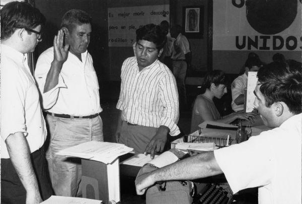 Jesus Salas, center, is the leader of Obreros Unidos (United Workers), a Chicano migrant workers' rights group started in the 1960s for the improvement of working conditions in Wisconsin's agricultural and manufacturing industries. To the immediate left of Salas is a farm worker giving an oath to be sworn in to file claims for workmen's compensation at the Obreros Unidos office. Salas is interpreting for volunteer lawyers Allen Samson (far left) and E. Michael Mc Cann (right). In the background to the right hangs a banner that says "Obreros Unidos," with the United Farm Worker's Aztec eagle symbol in the circle. At the center background is a portrait of Our Lady of Guadalupe. On the background to the left is a sign that says in English: "It is better to die barefoot but standing."<p>Trabajador agrícola da su juramento<p>Jesús Salas, en el centro, es el líder de Obreros Unidos, un grupo chicano a favor de los derechos de los trabajadores emigrantes que empezó en los años 1960 para mejorar las condiciones laborales en las industrias agrícolas y manufactureras en Wisconsin. Inmediatamente a la izquierda de Salas está un obrero agrícola dando su juramento para poder presentar reclamos de compensación de trabajadores en la oficina de Obreros Unidos. Salas está interpretando a los licenciados voluntarios, Allen Samson (al extremo izquierdo) y E. Michael Mc Cann (a la derecha). Al fondo y a la derecha cuelga un estandarte que dice "Obreros Unidos" y el símbolo de una águila Azteca de United Farm Workers en un círculo. Al fondo y al centro está un retrato de Nuestra Señora de Guadalupe. Al fondo hacia la izquierda está un letrero que dice "Es mejor morir descalzo pero en pie."</p>