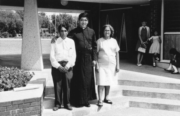 At the center stands Father Antonio Gonzales, a former migrant worker from Rio Grande City, Texas, who is visiting his parents in Wautoma. In 1966, he led a march for better working conditions in Texas. He has his arm around his father who is standing to the left with his hands folded in front of him. To the right is Father Gonzalez's mother wearing a dress and glasses. In the background to the right is a group of girls and a young woman wearing dresses and sunglasses.<p>This photograph is a part of Wisconsin-native David Giffey's series "Struggle for Justice," images from the migrant farm worker struggle including an independent organizing effort in Wisconsin and the nationwide grape boycott movement started by Cesar Chavez of United Farm Workers during the 1960s and 1970s.<p>El Padre González en Wautoma<p>En el centro está el Padre Antonio Gonzales, él anteriormente fue un trabajador emigrante de Rio Grande City, Texas, visitando a sus padres en Wautoma. En 1966, él guio una marcha para mejorar las condiciones laborales en Texas. Él tiene su brazo alrededor de su padre que está parado a su izquierda y con las manos dobladas enfrente de él. A la derecha del Padre González está su madre en un vestido y con anteojos. Al fondo y hacia la derecha está un grupo de niñas y una mujer joven en un vestido y con lentes de sol. Esta fotografía es parte de la serie "Lucha por la Justicia" tomada por David Giffey, originario de Wisconsin, las imágenes muestran la lucha de los trabajadores agrícolas emigrantes incluyendo un esfuerzo independiente organizado en Wisconsin y el movimiento nacional del boicot de uvas empezado por Cesar Chávez de la unión de campesinos o United Farm Workers durante los años 1960 y 1970.</p>