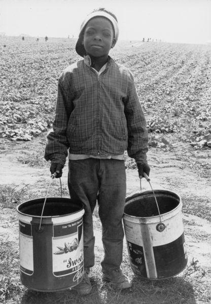 A nine-year old Jamaican boy wearing a backwards cap, plaid jacket, jeans, and sneakers. He is holding two buckets and is looking at the camera. He is joining a strike against Libby, McNeill & Libby, Inc. Behind him are other migrant farm laborers working in the fields of Central Wisconsin.<p>In 1942-1964, more than 4 million Mexican workers entered the U.S. for temporary employment under the Bracero Program which was an agreement between the U.S. and Mexico. The agreement was created in response to a lobby from growers in western states who anticipated labor shortages during World War II and the Korean War. Wisconsin's migrant labor force included workers from Jamaica, Barbados, Bahama, Honduras and prisoners of war from Germany and Italy.<p>This photograph is a part of Wisconsin-native David Giffey's series "Struggle for Justice," images from the migrant farm worker struggle including an independent oganizing effort in Wisconsin and
the nationwide grape boycott movement started by Cesar Chavez of United Farm Workers during the 1960s and 1970s. Many migrant farm laborers traveled from Texas to Wisconsin in search of seasonal field work.<p>Un niño hace huelga contra Libby, McNeill & Libby, Inc.<p>Un niño jamaiquino de 9 años de edad, vistiendo una gorra hacia atrás, una chaqueta con cuadros, un pantalón de mezclilla, y unos zapatos estilo tenis. Él está uniéndose a una huelga contra Libby, McNeill & Libby, Inc. Detrás de él hay otros obreros agrícolas emigrantes trabajando en los campos del centro de Wisconsin. Entre 1942-1964, más de 4 millones de trabajadores mexicanos entraron a los Estados Unidos para trabajar temporalmente bajo el programa de Braceros, el cual era un acuerdo entre los Estados Unidos y México. El acuerdo fue creado a causa de presión de parte de productores de los estados del oeste del país que anticipaban una escasez de trabajadores durante la Segunda Guerra Mundial y la guerra en Corea. La fuerza laboral emigrante de Wisconsin incluía trabajadores de Jamaica, Barbados, Bahamas, Honduras, y prisioneros de guerra de Alemania e Italia. Esta fotografía es parte de la serie "Lucha por la Justicia" tomada por David Giffey, originario de Wisconsin, las imágenes muestran la lucha de los trabajadores agrícolas emigrantes incluyendo un esfuerzo independiente organizado en Wisconsin y el movimiento nacional del boicot de uvas empezado por Cesar Chávez de la unión de campesinos o United Farm Workers durante los años 1960 y 1970. Muchos obreros agrícolas emigrantes viajaban desde Texas hacia Wisconsin en busca de trabajo temporal en los campos.</p?