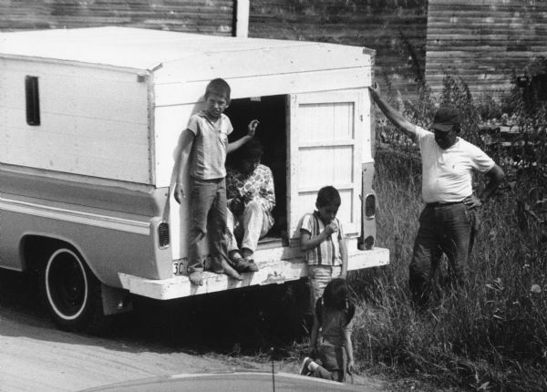 A family from Texas temporarily settles in Waushara County. Their white truck is parked in front of a grassy area and wooden house. The father is standing to the right, wearing a cap, white shirt and jeans, leaning on the back of the truck with one arm on the truck and the other on his hip. He is looking at his young daughter who has her back to the camera and is wearing pigtails and holding a stick. The rest of the family is in front or inside the truck. A shoeless boy stands on the edge of the back of the truck looking into the camera.<p>This photograph is a part of Wisconsin-native David Giffey's series "Struggle for Justice," images from the migrant farm worker struggle including an independent organizing effort in Wisconsin and the nationwide grape boycott movement started by Cesar Chavez of United Farm Workers during the 1960s and 1970s. Many migrant farm laborers traveled from Texas to Wisconsin in search of seasonal field work.<p>Familia emigrante de Texas<p>Una familia de Texas temporalmente se establece en el condado de Waushara. Su camioneta blanca está estacionada enfrente de una área con pasto y una casa de madera. El padre está parado a la derecha, vistiendo una gorra, camisa blanca, y pantalón de mezclilla, inclinándose en la parte trasera de la camioneta con un brazo en la camioneta y el otro en sus caderas. Está mirando a su hija que tiene su espalda hacia la cámara, con dos trenzas a los lados, y deteniendo un palo. El resto de la familia está enfrente o adentro de la camioneta. Un niño sin zapatos está parado en la parte trasera de la camioneta y mirando hacia la cámara. Esta fotografía es parte de la serie "Lucha por la Justicia" tomada por David Giffey, originario de Wisconsin, las imágenes muestran la lucha de los trabajadores agrícolas emigrantes incluyendo un esfuerzo independiente organizado en Wisconsin y el movimiento nacional del boicot de uvas empezado por Cesar Chávez de la unión de campesinos o United Farm Workers durante los años 1960 y 1970.</p>