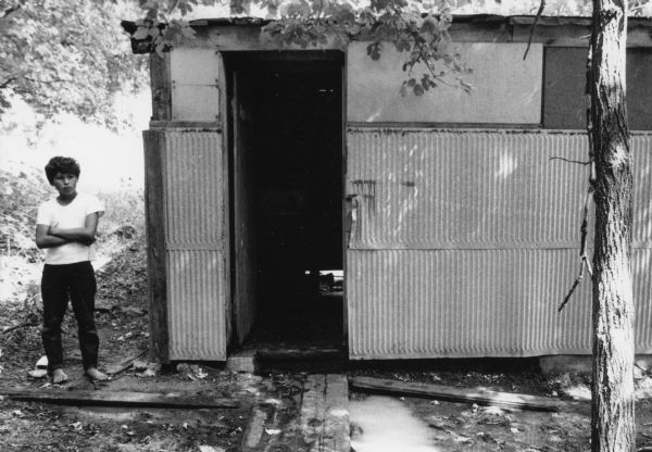 A boy wearing a white short-sleeved t-shirt and dark pants is barefoot and standing to the left of a communal shower hut for migrant farm workers in a Wautoma labor camp. The communal hut is floorless and has a tin exterior. Inside, there are shower heads and sinks.<p>This photograph is a part of Wisconsin-native David Giffey's series "Struggle for Justice," images from the migrant farm worker struggle including an independent organizing effort in Wisconsin and
the nationwide grape boycott movement started by Cesar Chavez of United Farm Workers during the 1960s and 1970s. Many migrant farm laborers traveled from Texas to Wisconsin in search of seasonal field work.<p>Baño comunal para trabajadores emigrantes<p>Un niño vistiendo una camiseta de manga corta, pantalón oscuro, y descalzado está parado hacia la izquierda de la choza que sirve como baño comunal para los trabajadores agrícolas en el campo laboral en Wautoma. La choza comunal no tiene piso y el exterior es de hojalata. Adentro hay unas regaderas (para ducharse) y unos lavamanos. Esta fotografía es parte de la serie "Lucha por la Justicia" tomada por David Giffey, originario de Wisconsin, las imágenes muestran la lucha de los trabajadores agrícolas emigrantes incluyendo un esfuerzo independiente organizado en Wisconsin y el movimiento nacional del boicot de uvas empezado por Cesar Chávez de la unión de campesinos o United Farm Workers durante los años 1960 y 1970. Muchos obreros agrícolas emigrantes viajaban desde Texas hacia Wisconsin en busca de trabajo temporal en los campos.</p>