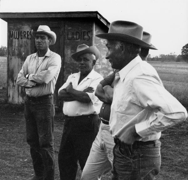 Migrant farm workers meeting in Bancroft after a strike. There are four men wearing hats standing in front of a women's outhouse. Strikes in Wisconsin were organized by the local farm-worker labor union, Obreros Unidos, an independent farm worker labor union effort in the 1960s.<p>Junta de trabajadores emigrantes<p>Trabajadores emigrantes en una junta después de una huelga en Bancroft, Wisconsin. Cuatro hombres con sombreros están parados enfrente de una letrina para mujeres. Las huelgas en Wisconsin fueron organizadas por el sindicato local de trabajadores agrícolas, Obreros Unidos, un esfuerzo independiente del sindicato de trabajadores agrícolas durante los años 1960.</p>