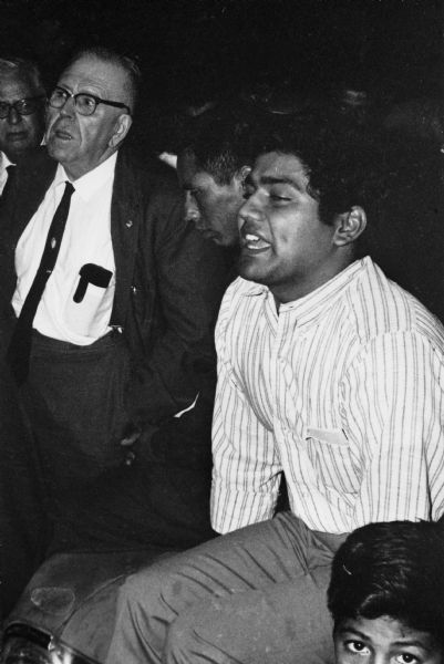 The man standing in a suit and dark tie with glasses to the left is Charles Heymanns, international organizer for AFL-CIO. Seated to his right in a light striped shirt is Jesus Salas, leader of Obreros Unidos (United Workers), Wisconsin's migrant farm workers' union. They are participating in a strike rally with other migrant workers, such as the man between Heymanns and Salas, and the boy in the bottom right corner.<p>Strikes in Wisconsin were organized by the Obreros Unidos, an independent farm worker labor union effort in the 1960s.<p>Charles Heymanns y Jesús Salas en una manifestación y huelga<p>El hombre parado a la izquierda y vistiendo un traje oscuro con corbata y con anteojos es Charles Heymanns, organizador internacional para AFL-CIO. Sentado a su derecha, vistiendo una camisa clara con rayas está Jesús Salas líder de Obreros Unidos, un sindicato para trabajadores agrícolas en Wisconsin. Ellos están participando en una manifestación y huelga con otros trabajadores emigrantes, por ejemplo, el hombre en medio de Heymanns y Salas, y el niño hacia el extremo derecho. Las huelgas en Wisconsin fueron organizadas por Obreros Unidos, un esfuerzo independiente del sindicato de trabajadores agrícolas durante los años 1960.</p>