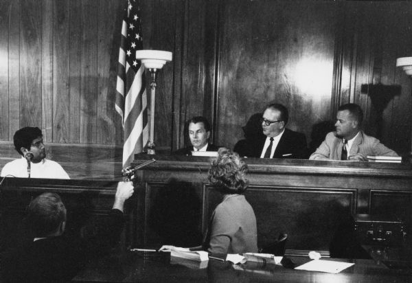 Migrant farm workers testify at the Wisconsin Employment Relations Commission (WERC) hearing after a strike in Bancroft. Seated on the left is Obreros Unidos (United Workers) migrant farm workers' union leader, Jesus Salas, testifying to the three men seated on an elevated bench to his right. In front of Salas is a man holding a microphone towards the bench to record the hearing. To his right is a woman with her head turned away from the camera and typing notes on a stenograph machine. There are papers scattered on the desk in front of her.<p>Strikes in Wisconsin were organized by the Obreros Unidos, an independent farm worker labor union effort in the 1960s.<p>Audiencia de WERC en Wautoma<p>Trabajadores agrícolas emigrantes testifican ante una audiencia con el comité de relaciones laborales o Wisconsin Employment Relations Commission (WERC) después de una huelga en Bancroft. Sentado a la derecha está el líder de Obreros Unidos, un sindicato para los derechos de los trabajadores agrícolas emigrantes, Jesús Salas, que está testificando ante los tres hombres sentados en una banca elevada hacia su derecha. Enfrente de Salas está un hombre deteniendo un micrófono hacia la banca para grabar la audiencia. Hacia su derecha está una mujer con su cabeza volteada alejada de la cámara y tomando notas en su máquina de estenografía. Hay papeles regados en el escritorio enfrente de ella. Las huelgas en Wisconsin fueron organizadas por Obreros Unidos, un esfuerzo independiente del sindicato de trabajadores agrícolas durante los años 1960.</p>