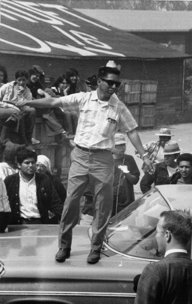 Standing on the hood of the car in a striped short-sleeved shirt and sunglasses is Salvador Sanchez, an organizer for Obreros Unidos (United Workers), a Wisconsin migrant farm workers' union. Behind Sanchez to his left is Jesus Salas, the leader of Obreros Unidos, wearing a black jacket. There is a group of female supporters seated in a row on a fence and a nun standing  behind Salas and Sanchez. The workers are preparing a strike.<p>Strikes in Wisconsin were organized by the Obreros Unidos, an independent farm worker labor union effort in the 1960s.<p>Salvador Sánchez se dirige a unos trabajadores<p>Parado en el cofre de un carro, vistiendo una camisa de manga corta y con lentes de sol está Salvador Sánchez, un organizador para Obreros Unidos, un sindicato de trabajadores agrícolas en Wisconsin. Detrás de Sánchez y a su derecha está Jesús Salas, líder de Obreros Unidos, vistiendo una chaqueta negra. Hay un grupo de mujeres simpatizantes sentadas en una cerca y una monja parada detrás de Salas y Sánchez. Los trabajadores están preparando una huelga. Las huelgas en Wisconsin fueron organizadas por Obreros Unidos, un esfuerzo independiente del sindicato de trabajadores agrícolas durante los años 1960.</p>