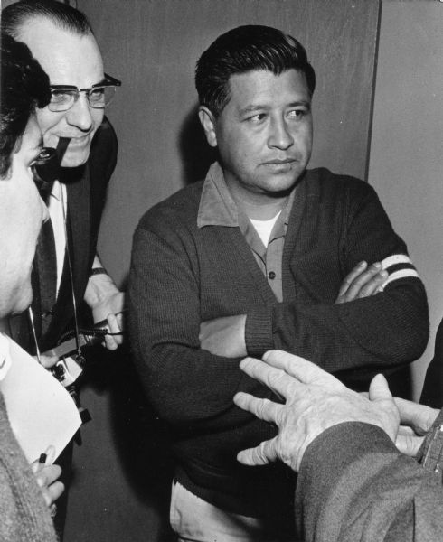 Cesar Chavez, the leader of United Farm Workers, is standing at the center of the photograph surrounded by members of the press. Behind him is a photographer with a pipe in his mouth and a camera in his hands. Besides him, someone is holding a pencil and paper taking notes. This is Chavez's first visit to Milwaukee to meet with Jesus Salas of Wisconsin's migrant farm worker union Obreros Unidos (United Workers) for El Centro Hispano-Americano to discuss agreement disputes between growers and farm workers<p>Strikes in Wisconsin were organized by the Obreros Unidos, an independent farm worker labor union effort in the 1960s.<p>Cesar Chávez en Wisconsin<p>Cesar Chávez líder de United Farm Workers, está parado en el centro de la fotografía rodeado de miembros de la prensa. Detrás de él está un fotógrafo con una pipa en su boca y una cámara en sus manos. A su lado, alguien está tomando notas con un lápiz y papel. Esta es la primera visita de Chávez a Milwaukee para conocer a Jesús Salas de Obreros Unidos, un sindicato de trabajadores agrícolas, para El Centro Hispano-Americano y para discutir acuerdos de disputa entre los agricultores y los trabajadores agrícolas. Las huelgas en Wisconsin fueron organizadas por Obreros Unidos, un esfuerzo independiente del sindicato de trabajadores agrícolas durante los años 1960.</p>