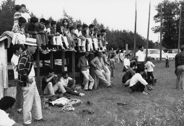 Obreros Unidos (United Workers) migrant farm worker union members at a Bancroft baseball game. Children sit on top of the dugout to watch. Adult men are standing and sitting on the grass. Cars are parked behind them.<p>This photograph is a part of Wisconsin-native David Giffey's series "Struggle for Justice," images from the migrant farm worker struggle including an independent oganizing effort in Wisconsin and the nationwide grape boycott movement started by Cesar Chavez of United Farm Workers during the 1960s and 1970s.<p>Juego de béisbol del sindicato de Bancroft<p>Unos trabajadores agrícolas emigrantes, miembros de Obreros Unidos en un juego de béisbol en Bancroft, Wisconsin. Unos niños están sentados arriba de un gallinero mientras miran. Unos hombres están parados y sentados en el pasto. Hay carros estacionados detrás de ellos. Esta fotografía es parte de la serie "Lucha por la Justicia" tomada por David Giffey, originario de Wisconsin, las imágenes muestran la lucha de los trabajadores agrícolas emigrantes incluyendo un esfuerzo independiente organizado en Wisconsin y el movimiento nacional del boicot de uvas empezado por Cesar Chávez de la unión de campesinos o United Farm Workers durante los años 1960 y 1970.</p>