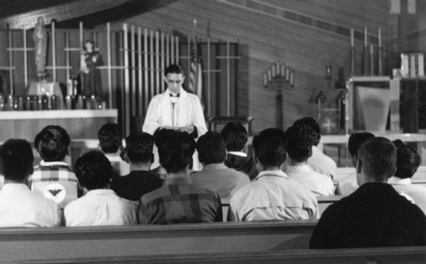 Father Michael Garrigan in the background wearing a white robe and glasses holding 8 a.m. mass at St. Joseph's Church with 30 marchers before a Obreros Unidos (United Workers) "march for respectability" beginning on Highway 21 and ending at the Capitol in Madison.<p>This photograph is a part of Wisconsin-native David Giffey's series "Struggle for Justice," images from the migrant farm worker struggle including an independent organizing effort in Wisconsin and the nationwide grape boycott movement started by Cesar Chavez of United Farm Workers during the 1960s and 1970s.<p>El Padre Garrigan oficiando una misa antes de una marcha<p>El Padre Michael Garrigan está hacia el fondo vestido en habito blanco y con lentes, oficiando una misa a las 8 a.m. en la iglesia St. Joseph's Church con 30 protestantes antes de una marcha de Obreros Unidos, "una marcha de respetabilidad" empezando en Highway 21 y terminando en el Capitolio en Madison. Esta fotografía es parte de la serie "Lucha por la Justicia" tomada por David Giffey, originario de Wisconsin, las imágenes muestran la lucha de los trabajadores agrícolas emigrantes incluyendo un esfuerzo independiente organizado en Wisconsin y el movimiento nacional del boicot de uvas empezado por Cesar Chávez de la unión de campesinos o United Farm Workers durante los años 1960 y 1970.</p>