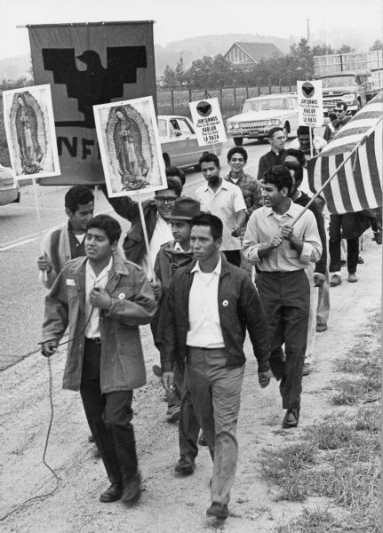 30 marchers of Obreros Unidos (United Workers) march from Wautoma to Madison on Highway 21 to petition lawmakers to hold farms and food industry corporations accountable for better working conditions for migrant farm workers. Jesus Salas, in front leading the march, is holding a microphone. Next to him is Salvador Sanchez, also a march leader. Two men behind Salas carry images of Our Lady of Guadalupe. One man in glasses is carrying the National Farm Workers Association banner with the Aztec eagle symbol. To the right, a man is holding the American flag. With Father Garrigan, in the middle of the march, are two men holding signs that say "Juntarnos para ser reconocidos. Hablar para ser oidos. La raza tiene causa"/ "Join us to be recognized. Speak to be heard. The race has cause."<p>Salas y Sánchez guían la marcha hacia Madison<p>30 manifestantes de Obreros Unidos marchan desde Wautoma hacia Madison en la carretera Highway 21 para peticionar a los legisladores estatales que mantengan responsables a los agricultores y corporaciones en la industria de comida para el mejoramiento de las condiciones de los trabajadores agrícolas emigrantes. Jesús Salas, enfrente y guiando la marcha, está sosteniendo un micrófono. A su lado está Salvador Sánchez, también un líder de sindicato. Dos hombres detrás de Salas cargan imágenes de Nuestra Señora de Guadalupe. Un hombre con anteojos está cargando el estandarte de National Farm Workers Association (NFWA) con el símbolo de una águila Azteca. A la derecha, un hombre está sosteniendo la banderea americana. Con el Padre Garrigan, en medio de la fila de marcha, están dos hombres con letreros que dicen, "Juntarnos para ser reconocidos. Hablar para ser oídos. La raza tiene causa." Esta fotografía es parte de la serie "Lucha por la Justicia" tomada por David Giffey, originario de Wisconsin, las imágenes muestran la lucha de los trabajadores agrícolas emigrantes incluyendo un esfuerzo independiente organizado en Wisconsin y el movimiento nacional del boicot de uvas empezado por Cesar Chávez de la unión de campesinos o United Farm Workers durante los años 1960 y 1970.<p>This photograph is a part of Wisconsin-native David Giffey's series "Struggle for Justice," images from the migrant farm worker struggle including an independent oganizing effort in Wisconsin and the nationwide grape boycott movement started by Cesar Chavez of United Farm Workers during the 1960s and 1970s.</p>