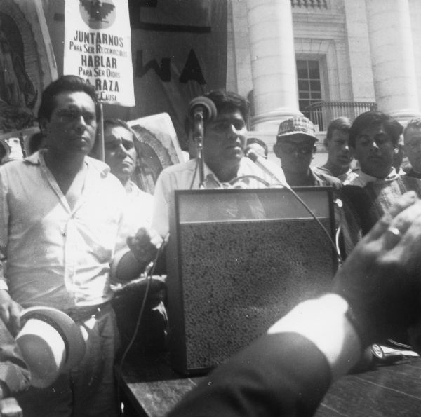 Jesus Salas addresses a rally at a podium in front of the Wisconsin State Capitol. Behind him are marchers who began the march from Wautoma and walked to Madison on Highway 21. Marchers are holding images of Our Lady of Guadalupe, a National Farm Worker Association's banner, and signs with the message "Juntarnos para ser reconocidos. Hablar para ser oidos. La raza tiene causa"/ "Join us to recognized. Speak to be heard. The people have a cause."<p>This photograph is a part of Wisconsin-native David Giffey's series "Struggle for Justice," images from the migrant farm worker struggle including an independent oganizing effort in Wisconsin and the nationwide grape boycott movement started by Cesar Chavez of United Farm Workers during the 1960s and 1970s.<p>Salas se dirige al público<p>Jesús Salas le habla al público desde un podio enfrente del Capitolio del estado de Wisconsin. Detrás de él están unos manifestantes que marcharon desde Wautoma y caminaron hacia Madison en la carretera Highway 21. Unos manifestantes están sosteniendo imágenes de Nuestra Señora de Guadalupe, el estandarte de National Farm Workers Association (NFWA), y letreros que dicen, "Juntarnos para ser reconocidos. Hablar para ser oídos. La raza tiene causa." Esta fotografía es parte de la serie "Lucha por la Justicia" tomada por David Giffey, originario de Wisconsin, las imágenes muestran la lucha de los trabajadores agrícolas emigrantes incluyendo un esfuerzo independiente organizado en Wisconsin y el movimiento nacional del boicot de uvas empezado por Cesar Chávez de la unión de campesinos o United Farm Workers durante los años 1960 y 1970.</p>