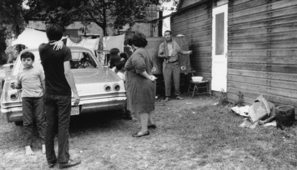 A migrant worker family stands in front of a parked vehicle and a cabin that serves as temporary housing in a Wautoma labor camp. Some of the children are not wearing shoes. The shirtless man with a towel around his neck is washing or shaving himself with water in a small basin beside him.<p>This photograph is a part of Wisconsin-native David Giffey's series "Struggle for Justice," images from the migrant farm worker struggle including an independent organizing effort in Wisconsin and the nationwide grape boycott movement started by Cesar Chavez of United Farm Workers during the 1960s and 1970s. Many migrant farm laborers traveled from Texas to Wisconsin in search of seasonal field work.<p>Vivienda para trabajadores emigrantes<p>Una familia de trabajadores emigrantes está parada enfrente de un vehículo estacionado y una cabaña que sirve como vivienda temporal en el campo laboral en Wautoma. Algunos de los niños no traen zapatos. El hombre sin camisa, con una toalla alrededor de su cuello, está lavándose o rasurándose con agua del contenedor a su lado. Esta fotografía es parte de la serie "Lucha por la Justicia" tomada por David Giffey, originario de Wisconsin, las imágenes muestran la lucha de los trabajadores agrícolas emigrantes incluyendo un esfuerzo independiente organizado en Wisconsin y el movimiento nacional del boicot de uvas empezado por Cesar Chávez de la unión de campesinos o United Farm Workers durante los años 1960 y 1970. Muchos obreros agrícolas emigrantes viajaban desde Texas hacia Wisconsin en busca de trabajo temporal en los campos.</p>