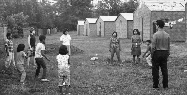 A group of children of migrant worker families playing a ball game in a circle with an adult who has his back to the camera at a Waushara County Labor Camp. Around them are buildings that serve as temporary housing for migrant farm workers.<p>This photograph is a part of Wisconsin-native David Giffey's series "Struggle for Justice," images from the migrant farm worker struggle including an independent organizing effort in Wisconsin and the nationwide grape boycott movement started by Cesar Chavez of United Farm Workers during the 1960s and 1970s. Many migrant farm laborers traveled from Texas to Wisconsin in search of seasonal field work.<p>Unos niños en el campo laboral en Waushara<p>En el campo laboral Waushara County Labor Camp, un grupo de niños de las familias de trabajadores emigrantes están jugando un juego de pelota en un círculo con un adulto que tiene su espalda hacia la cámara. A su alrededor hay edificios que servían como viviendas temporales para los trabajadores agrícolas emigrantes. Esta fotografía es parte de la serie "Lucha por la Justicia" tomada por David Giffey, originario de Wisconsin, las imágenes muestran la lucha de los trabajadores agrícolas emigrantes incluyendo un esfuerzo independiente organizado en Wisconsin y el movimiento nacional del boicot de uvas empezado por Cesar Chávez de la unión de campesinos o United Farm Workers durante los años 1960 y 1970. Muchos obreros agrícolas emigrantes viajaban desde Texas hacia Wisconsin en busca de trabajo temporal en los campos.</p>