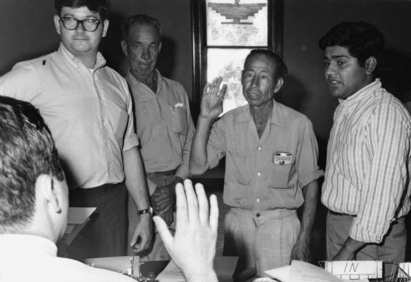 Jesus Salas, right, is the leader of Obreros Unidos (United Workers), a Chicano migrant workers' rights group started in the 1960s for the improvement of working conditions in Wisconsin's agricultural and manufacturing industries. To the immediate left of Salas is a farm worker with his arm raised giving an oath to be sworn in to file claims for workmen's compensation. Salas is interpreting for volunteer lawyers Allen Samson (far left) and E. Michael Mc Cann (back to camera sitting at desk). The man to the right of Allen Samson is unidentified. In the background is a window decorated with the United Farm Worker's Aztec eagle symbol.<p>Salas interpretando a favor de la compensación de los trabajadores<p>Jesús Salas, a la derecha, es el líder de Obreros Unidos, un grupo chicano a favor de los derechos de los trabajadores emigrantes que empezó en los años 1960 para mejorar las condiciones laborales en las industrias agrícolas y manufactureras en Wisconsin. Inmediatamente a la izquierda de Salas está un trabajador agrícola con su brazo levantado mientras da su juramento para poder presentar reclamos de compensación de trabajadores. Salas está interpretando por los licenciados voluntarios, Allen Samson (al extremo izquierdo) y E. Michael Mc Cann (sentado, con su espalda hacia la cámara). El hombre a la derecha de Allen Samson no está identificado. Hacia el fondo hay una ventana decorada con el símbolo de una águila Azteca como la imagen usada por United Farm Workers.</p>