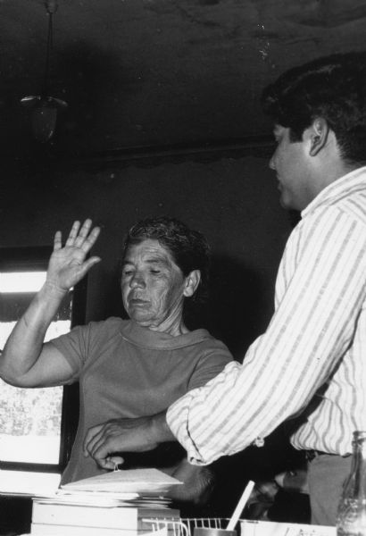 Jesus Salas, right, is the leader of Obreros Unidos (United Workers), a Chicano migrant workers' rights group started in the 1960s for the improvement of working conditions in Wisconsin's agricultural and manufacturing industries. To the immediate left of Salas is a farm worker giving an oath to be sworn in to file claims for workmen's compensation at the Obreros Unidos office.<p>Trabajador agrícola de edad avanzada tomando su juramento<p>Jesús Salas, a la derecha, es el líder de Obreros Unidos, un grupo chicano a favor de los derechos de los trabajadores emigrantes que empezó en los años 1960 para mejorar las condiciones laborales en las industrias agrícolas y manufactureras en Wisconsin. Inmediatamente a la izquierda de Salas está un obrero agrícola dando su juramento para poder presentar reclamos de compensación de trabajadores en la oficina de Obreros Unidos.</p>