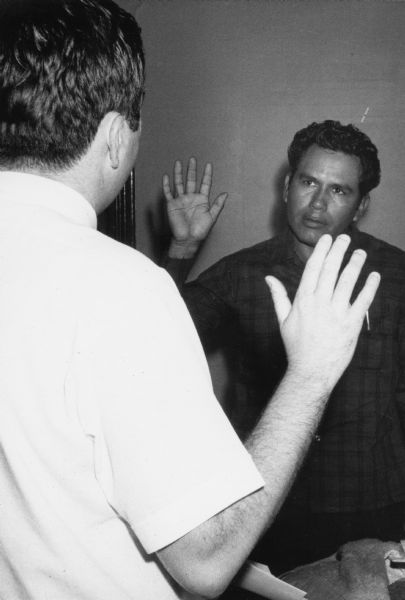 A farm worker, facing camera, is sworn in before filing a claim for worker’s compensation at the Obreros Unidos union office in Wautoma in 1967. A volunteer union lawyer, back to camera, is administering the oath. The worker’s compensation claims filed by Obreros Unidos members were the first such claims filed in Wisconsin on behalf of migrant farm workers.<p>Toma de juramento de un trabajador agrícola<p>Un obrero agrícola en presencia de un voluntario está dando su juramento para poder presentar reclamos de compensación de trabajadores en la oficina de Obreros Unidos. Obreros Unidos, es un grupo chicano a favor de los derechos de los trabajadores emigrantes que empezó en los años 1960 para mejorar las condiciones laborales en las industrias agrícolas y manufactureras en Wisconsin.</p>