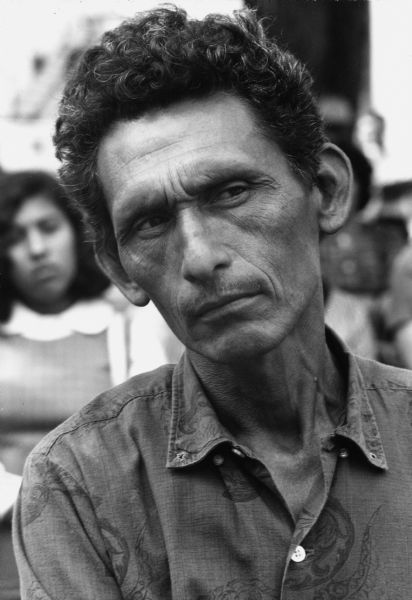 Close-up portrait of a migrant farm worker listening to speeches at an Oberos Unidos (United Workers) pre-march rally at Waushara Courthouse. The march was led by Jesus Salas to express the demands of migrant farm workers to provide them with $1.25 minimum wage, insurance benefits, a meeting with the governor's committee, and public sanitation facilities in Wautoma.<p>Strikes, rallies and marches in Wisconsin were organized by the Obreros Unidos, an independent farm worker labor union effort in the 1960s.<p>Trabajador emigrante en la manifestación en Wautoma<p>Retrato de cerca de un trabajador agrícola emigrante mientras escucha los discursos antes de la marcha de Obreros Unidos en el tribunal o Waushara Courthouse. La marcha fue dirigida por Jesús Salas para expresar las demandas de los trabajadores agrícolas emigrantes, incluyendo un sueldo mínimo de $1.25 por hora, beneficios de aseguranza, una junta con el comité del gobernador, e instalaciones sanitarias públicas para los trabajadores en Wautoma. Las huelgas, manifestaciones, y marchas en Wisconsin fueron organizadas por Obreros Unidos, un esfuerzo independiente del sindicato de trabajadores agrícolas durante los años 1960.</p>